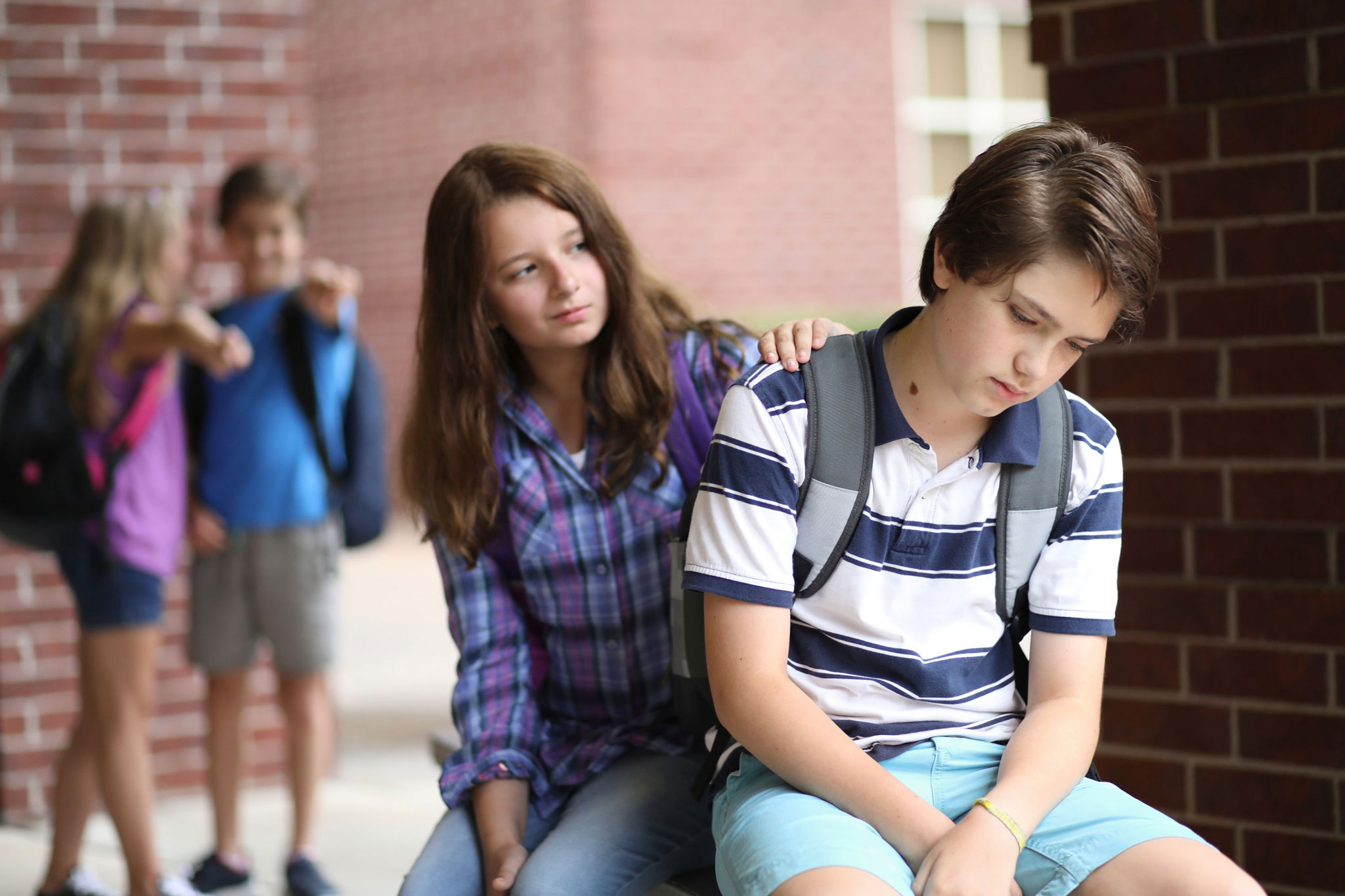 A boy sits looking upset on a school bench while a classmate rests a comforting hand on his shoulder; in the blurred background, two peers point toward him
