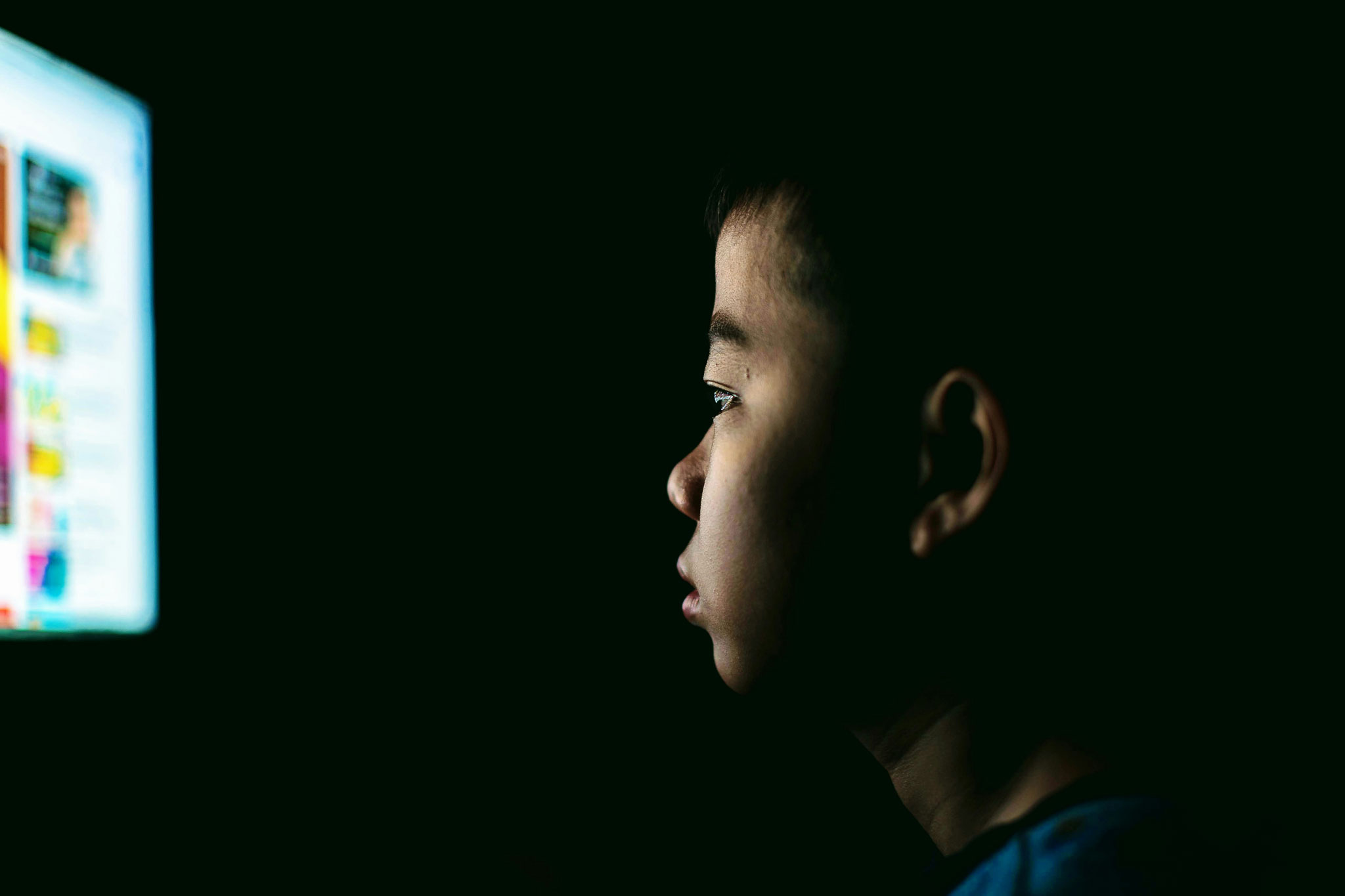 A young boy in profile looking intently at a glowing computer screen in a dark room, illustrating a child's focus on digital content